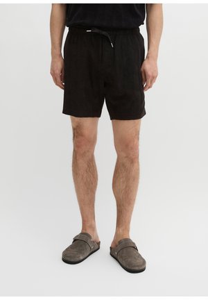 Mężczyzna w czarnych szortach ze ściągaczem i szarych butach typu slip-on stoi na jednolitym jasnym tle.