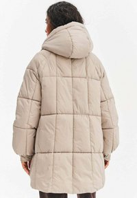 Béžová péřová bunda s kapucí, prošívaným vzorem a oversized rukávy. Textura vypadá měkká a vycpaná pro teplo.