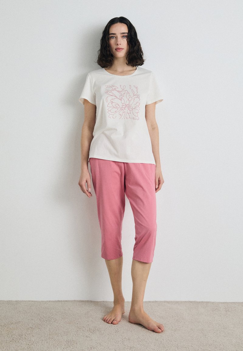 T-shirt a maniche corte in cotone crema con design floreale in contorno rosa, abbinato a pantaloni in cotone rosa a metà polpaccio. La modella è a piedi nudi.