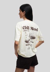 Cremefarvet t-shirt med en brun grafisk tryk af cocktails, tekst der siger "Chill Mood", og yderligere dekorative elementer.