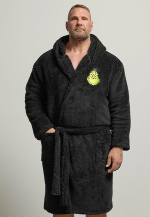 Homme portant un peignoir noir duveteux avec capuche et une broderie jaune du visage du Grinch sur la poitrine, debout devant un fond uni.