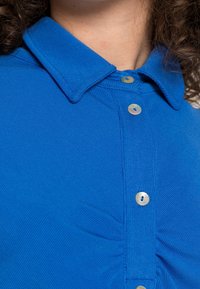 Sininen kauluspaita, jossa nappisuljenta, ribbineulospinta ja hienovarainen laskostus yksityiskohta liepeen läheisyydessä. Napit ovat helmiäishohtoiset.