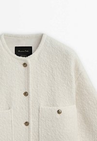 Kremowa kurtka z wełny z dodatkiem innych włókien, o teksturowanej powierzchni, okrągłym dekolcie, trzech metalowych guzikach oraz dwóch przednich kieszeniach. Widoczna metka marki wewnątrz.