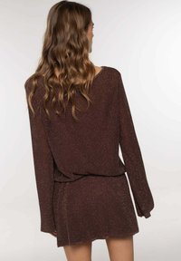 Vestido de punto marrón con mangas largas, que presenta un corte holgado, una superficie texturizada y un sutil efecto brillante. Largo corto con cinturilla elástica.