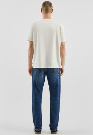 Man in een effen wit T-shirt met korte mouwen en een blauwe jeans, staand met zijn rug naar de camera tegen een effen lichte achtergrond.