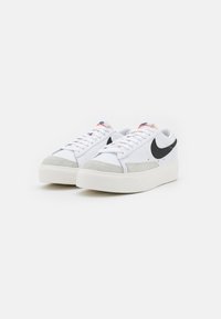 Zapatillas Nike de corte bajo en blanco con logotipos de swoosh en negro, acentos de ante gris en la puntera y suelas gruesas blancas, inclinadas de lado sobre un fondo blanco.