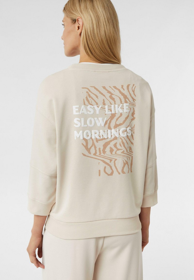 Donna bionda che indossa un felpa beige con motivo zebrastripe beige e testo bianco "FACILE COME LE LENTE MATTINE" sulla schiena.