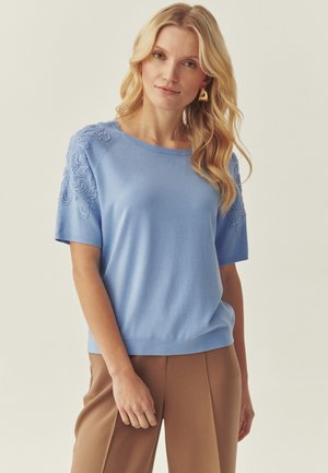 Frau mit langen blonden Haaren, die goldene Ohrringe, ein hellblaues Kurzarmoberteil mit strukturierten Schultern und braune Hosen trägt.