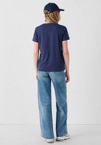 Una donna di spalle indossa un cappellino blu navy, una maglietta blu navy a maniche corte, jeans a gamba larga di un azzurro chiaro e sneakers bianche su uno sfondo bianco neutro.