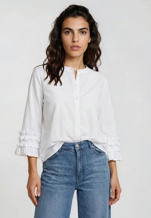 Frau mit welligem dunklem Haar, die eine weiße, geknöpfte Bluse mit gerafften Dreiviertelärmeln und blaue Jeans mit hoher Taille trägt.
