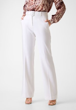 Frau trägt weiße, hochtaillierte, weit geschnittene Hose mit Gürtel, kombiniert mit einer gemusterten, langärmligen Bluse und goldfarbenen Sandalen mit offenem Zehenbereich.