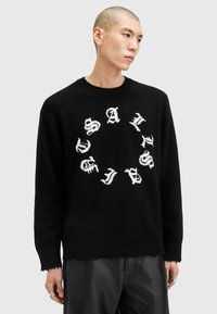 Sort strikket sweater med et cirkulært design og hvid tekst, rund halsudskæring og frynset kant. Materialet ser blødt ud med en tekstureret finish.