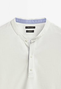 Vit avslappnad polo-skjorta i en strukturerad tygkvalitet, med bandkrage och två-knapps plackett; har en ljusblå accent på insidan av kragen.