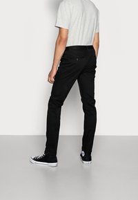 Pantaloni in cotone neri con vestibilità tapered, tasche posteriori visibili e una texture liscia. Abbinati a sneakers nere e una t-shirt grigia.
