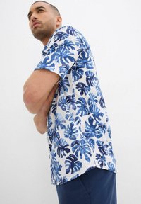 Camicia a maniche corte con sfondo bianco, decorata con motivi di foglie di monstera blu. Realizzata in tessuto leggero, con una vestibilità comoda.