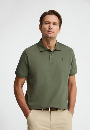 Mann mit kurzen braunen Haaren, der ein olivgrünes Polohemd mit kurzen Ärmeln und beige Hosen trägt, steht mit den Händen in den Taschen.