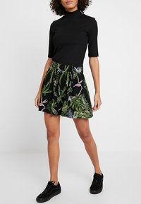 Haut noir à col roulé côtelé associé à une jupe noire ornée de motifs de feuilles tropicales vertes. La tenue est complétée par des baskets noires.