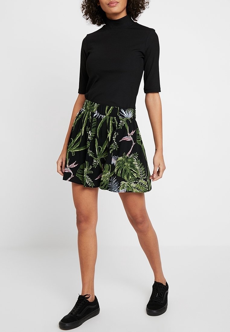 Haut noir à col roulé côtelé associé à une jupe noire ornée de motifs de feuilles tropicales vertes. La tenue est complétée par des baskets noires.