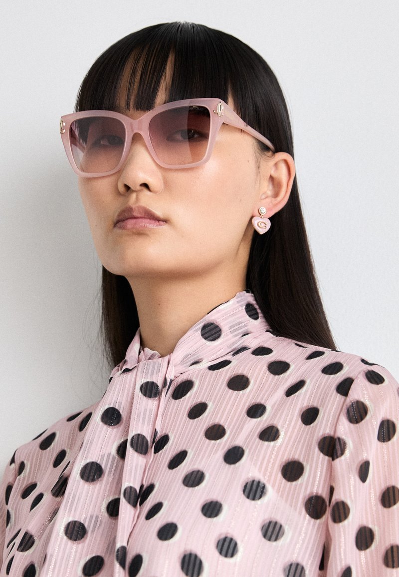 Różowe, oversized okulary przeciwsłoneczne z gradientowymi soczewkami, przezroczystymi oprawkami i złotymi detalami. Noszone z różową bluzką w kropki z plisami.