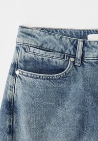 Sezione di jeans in denim blu chiaro che mostra la cintura, il passante per cintura, la tasca anteriore e la tasca portamonete con cuciture visibili e rivetti in metallo.