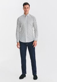 Biała koszula na guziki w czarne kropki, dopasowany krój, zestawiona z ciemnymi dżinsami i czarnymi butami, stojąca na białym tle.