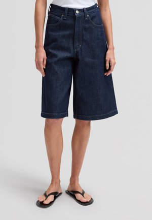 Sötétkék farmer térd alatti rövidnadrágot, fehér felsőt és fekete papucsot viselő személy világos háttér előtt áll.