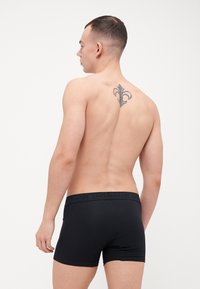 Sorte boxer briefs laget av glatt stoff, med en elastisk midjebånd med merkevarenavnet. Modellen viser en sidevisning som viser en tatovering.