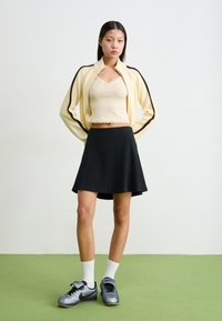 Kremni zip-up kardigan z temnimi črtami, bež majica na naramnice, črna plisirana krila in sivi Nike superge z belimi nogavicami, stoji na zeleni tleh.