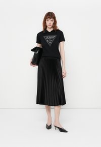 Black T-shirt with a triangular rhinestone logo, paired with a black pleated midi skirt and black pointed-toe heels, holding a black clutch.
