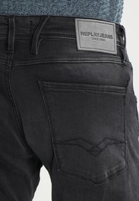 Jeans de mezclilla negra con un parche de cuero etiquetado "REPLAY JEANS" y un diseño de costura en zigzag sutil en el bolsillo trasero.