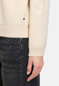 Cremfarbener Pullover mit einem strukturierten Muster an den Ärmeln, kombiniert mit dunkelgrauen Jeans, die seitlich verstreute Pailletten aufweisen.