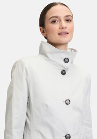 Lichtgrijze, waterdichte jas met hoge kraag, voorzien van drie grote knopen en een subtiele textuur. Slim fit, minimalistisch ontwerp.