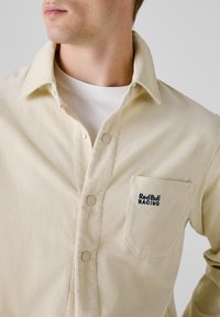 Beige corduroy shirt met een knoopsluiting, voorzien van een borstzak en het "Red Bull Racing" logo in marineblauw, met de benadrukking van ribbeltextuur.
