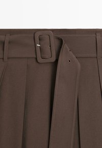 Hnědá sukně s vysokým pasem s širokým látkovým páskem provlečeným poutky na opasek a zajištěným obdélníkovou sponou a kovovým hrotem.