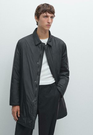 Black lightweight coat with a collared neckline, snap buttons, and long sleeves. Worn over a white sweater and black trousers.