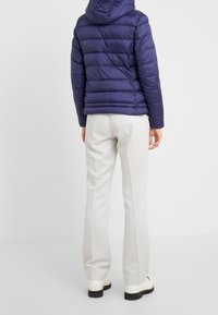 Giubbotto imbottito blu navy con cuciture orizzontali e cappuccio, abbinato a pantaloni grigio chiaro e stivali con suola bianca spessa, visto da dietro.