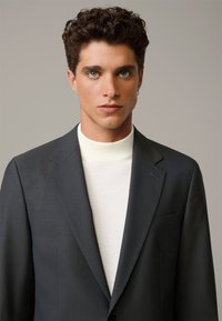 Giacca di suit grigio scuro con un sottile motivo a quadri, abbinata a un maglione a collo alto crema. Vestibilità liscia e sartoriale.