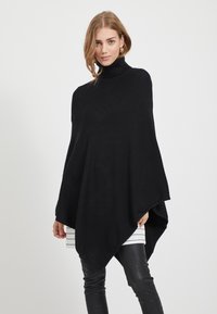 Poncho preto de gola alta, feito de um tecido knit suave, com uma silhueta drapeada, bainha pontiaguda e aberturas largas para os braços.