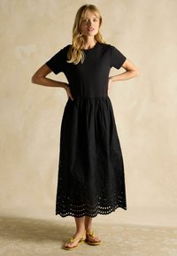 Melna kleita ar īsām piedurknēm un pieguļošu krūšu daļu, ar plūstošu svārku, kas rotāts ar acs izšuvumu un izgriezumiem. Valkāta ar dzeltenām sandālēm.