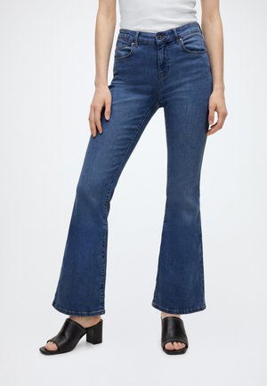Vrouw draagt donkerblauwe flare jeans met hoge taille en zwarte open-toe sandalen met hak, staand tegen een effen lichte achtergrond.