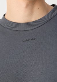 Felpa grigia girocollo con collo a costine, caratterizzata da un logo nero "Calvin Klein" stampato sul davanti. Texture liscia e design aderente.