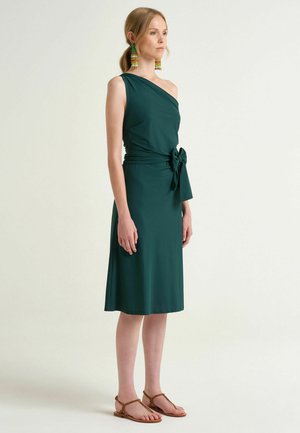 Donna che indossa un abito verde scuro monospalla con un fiocco in vita, orecchini colorati pendenti e sandali piatti marroni, in piedi contro uno sfondo semplice.