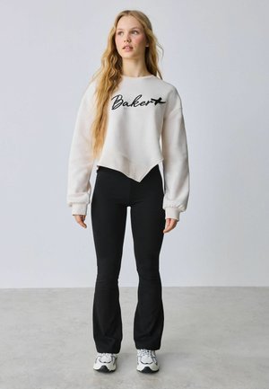 Ung kvinde med langt bølget hår iført en hvid "Baker" sweatshirt, sorte vidde bukser og hvide sneakers, stående på en grå gulv.