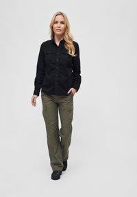 Camicia nera a bottoni con due tasche sul petto, abbinata a pantaloni cargo verde oliva. Modello indossa scarpe nere, in piedi contro uno sfondo grigio.