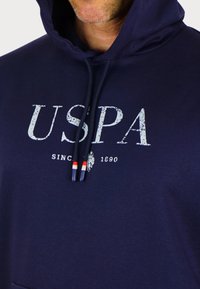 Sötétkék kapucnis pulóver, amely pamutból készült, nagy, megkopott fehér "USPA" felirattal. Piros és kék végű húzózsinórok tartoznak hozzá.