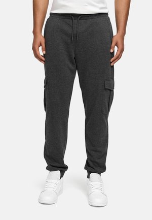 Man draagt donkergrijze cargojoggingbroek met trekkoord in de taille en zijdelingse klepzakken, gecombineerd met witte sneakers en een wit overhemd.