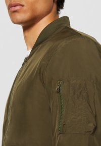 Olivově zelená bomber bunda s hladkým povrchem, žebrovaným límcem a bočními kapsami se zipy pro bezpečné zavření.