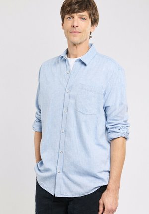 Homme portant une chemise bleu clair à boutons avec les manches retroussées et un pantalon noir, debout devant un fond clair uni.