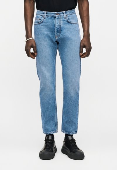 Personne portant un jean droit bleu clair avec un léger délavage, des chaussettes noires, des baskets noires et une chemise noire, debout devant un fond blanc.