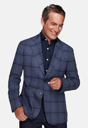 Man smiling while buttoning a blue plaid blazer over a dark navy shirt and dark jeans against a plain white background.